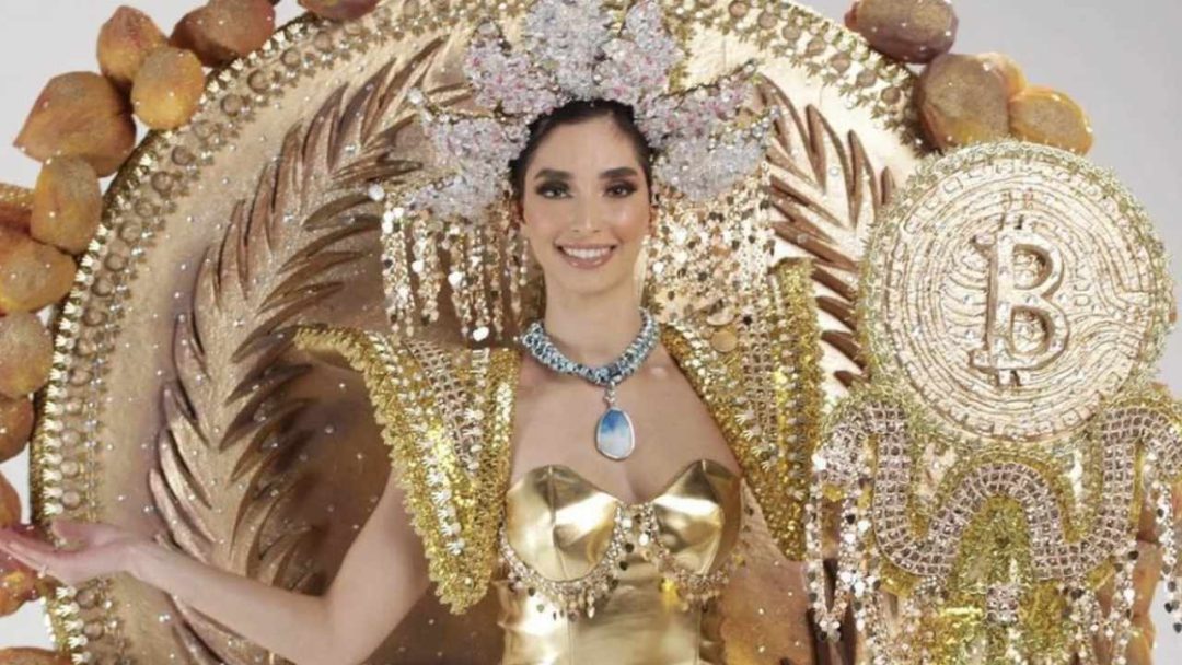 Foto da Miss El Salvador vestindo seu traje temático, segurando um cajado com o símbolo do Bitcoin.